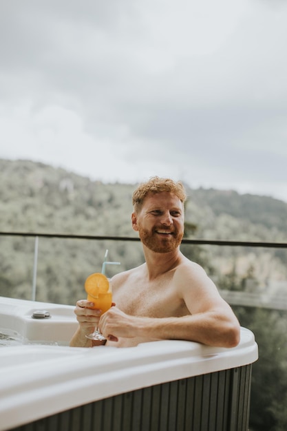 Young man enjoying in outdoor hot tub and drink fresh orange juice on vacation