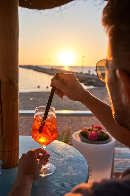 Young man enjoying and relaxing drinking a fresh spritz watching the sunset at a beach bar