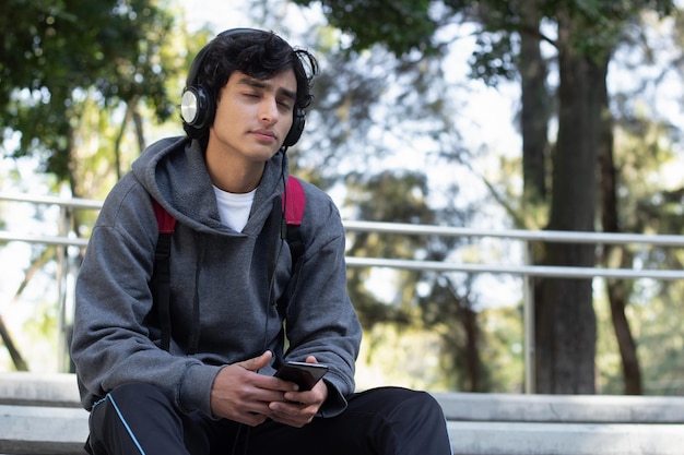 Young man enjoying some time alone relaxing listening to music with his headphones in the park, he has a smile on his face and a facial expression of peace and well-being.