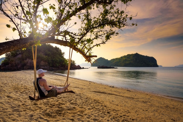 Young man enjoys sunset on a swing at a beach in thailand