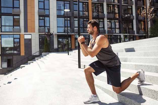 Young man exercising outside. Side view of sporty athletist down downhill exercise squat on one leg. Training outside at urban building. Hands together and concentration on exercise.