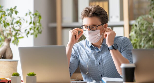 Young man in face mask is working on a laptop in office.