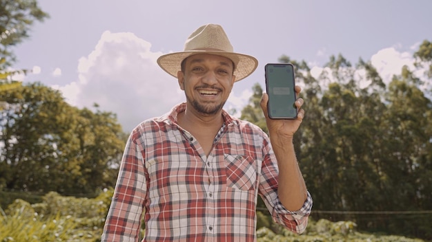 Young man farmer in hat smiling to camera and showing smartphone with chroma key screen