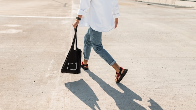 Young man in fashionable summer clothes in stylish red sandals with a black vintage bag runs down the street.