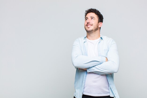 Young man feeling happy, proud and hopeful, wondering or thinking, looking up to copy space with crossed arms