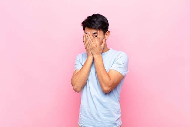 Young man feeling scared or embarrassed, peeking or spying with eyes half-covered with hands against flat color wall