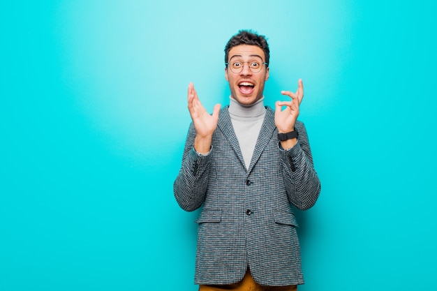 Young man feeling shocked and excited, laughing, amazed and happy because of an unexpected surprise against blue wall