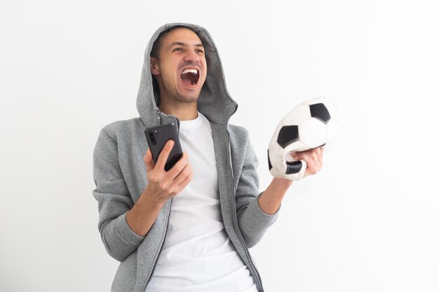 Young man football fan in shirt support team with soccer ball sit sofa home watch tv use mobile cell phone blank screen workspace area show thumb up isolated on orange background People sport concept.