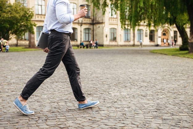 young man in formal wear, walking through city street holding takeaway coffee and silver laptop