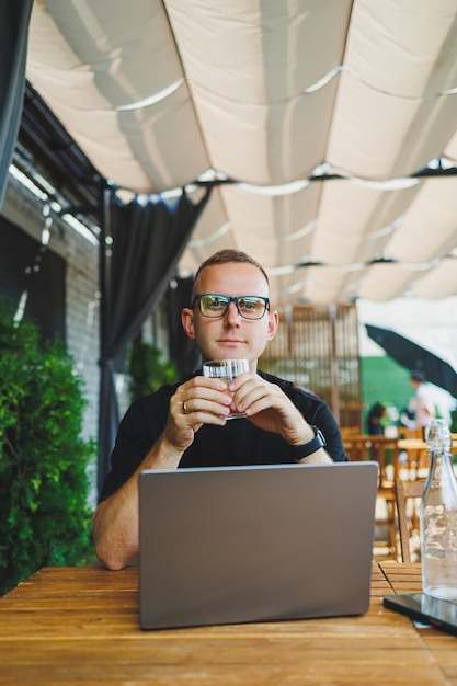 Young man freelancer developer enjoying coffee break on summer terrace of cozy cafe working remotely on laptop smiling looking at laptop