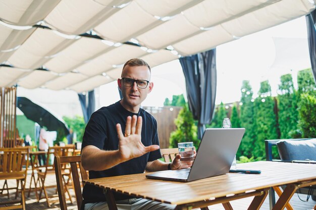 Young man freelancer developer enjoying coffee break on summer terrace of cozy cafe working remotely on laptop smiling looking at laptop