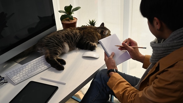 Young man freelancer making note on empty notebook while sitting with his cat at home.