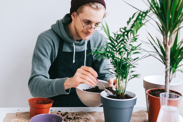 Young man gardener in eyeglasses transplanting plant in pots on the white wooden table and using tablet computer