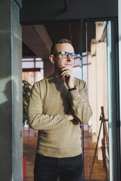 A young man in glasses and casual clothes stands in a modern workspace Male freelancer works remotely