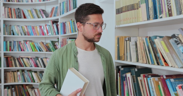 Young man in glasses and green shirt with a book in his hand walks near the shelves with book in the library University library concept