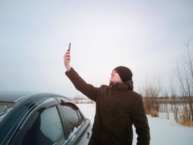 A young man got lost in a car in winter and is trying to catch the mobile network after getting out of the car. Concept of winter road travel, road search, navigation.