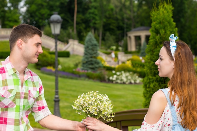 Young man in green and red plaid shirt gives a girl a bouquet of little white flowers on footbridge in park