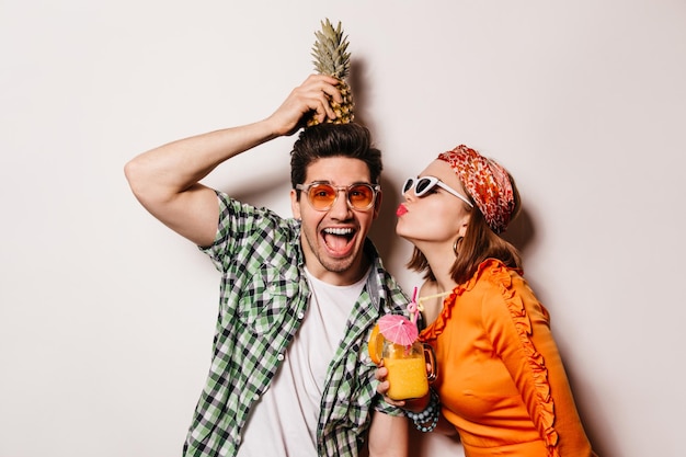 Young man in green shirt and white tshirt holds pineapple on his head Girl in orange dress with cocktail in her hands kisses man on cheek