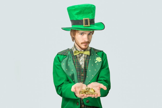 Young man in green suit hold golden coins in hands. He look  seriously. Guy wear saint Patrick's suit. Isolated on grey .