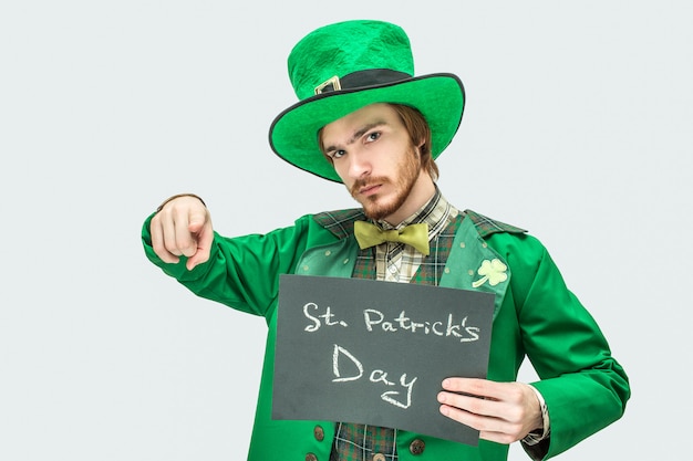 Young man in green suit holding dark tablet with St. Patrick's writing on it. He point straight and look . Isolated on grey .