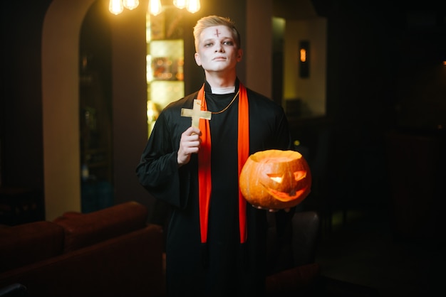 Young man in halloween priest costume holds carved pumpkin and religious cross