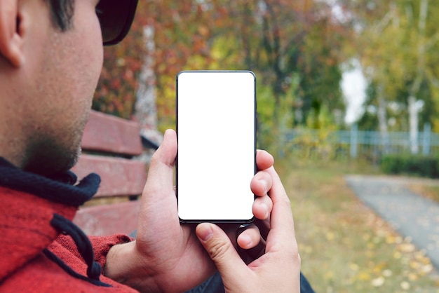 Young man hand holding smartphone with white screen outdoors against autumn sidewalk guy looks at the screen of an empty smartphone in daylight use smartphone outdoors in the open air