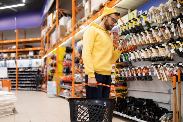 A young man in a hardware store next to a rack with hammers and axes looks at the smartphone screen