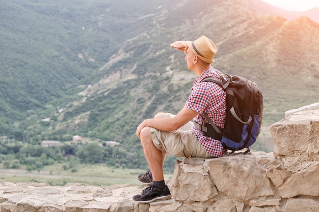 Young man in a hat with a backpack sits on a stone wall against a background of green mountains and looks into the distance