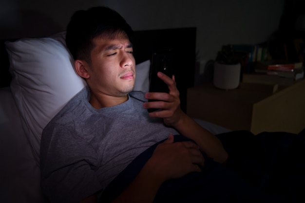 Young man having sore and tired eyes when using smartphone while lying in bed at night