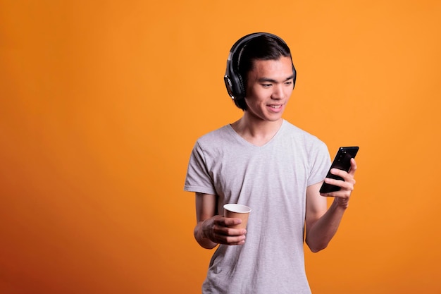 Young man in headphones watching video on mobile phone, holding coffee paper cup. Smiling asian teenager, using social media on smartphone, enjoying music, drinking take out tea