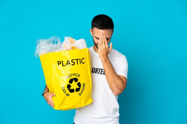 Young man holding a bag full of plastic bottles to recycle isolated on blue wall with tired and sick expression