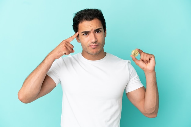 Young man holding a Bitcoin over isolated blue background having doubts and thinking