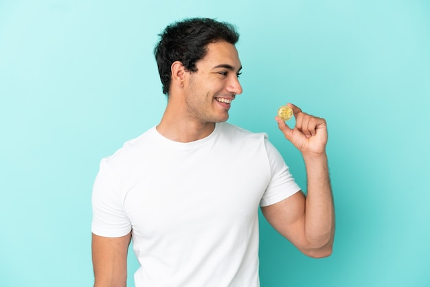 Young man holding a Bitcoin over isolated blue background looking side