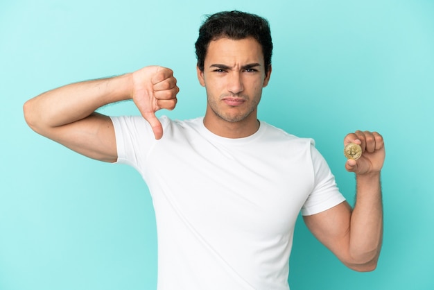 Young man holding a Bitcoin over isolated blue background showing thumb down with negative expression