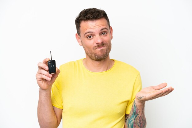 Young man holding car keys isolated on white background making doubts gesture while lifting the shoulders