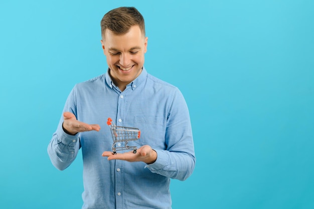 Photo a young man holding a mini shopping cart and standing in front of a blue background in the studio. sales concept. place for text.