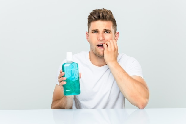 Young man holding a mouthwash biting fingernails, nervous and very anxious.