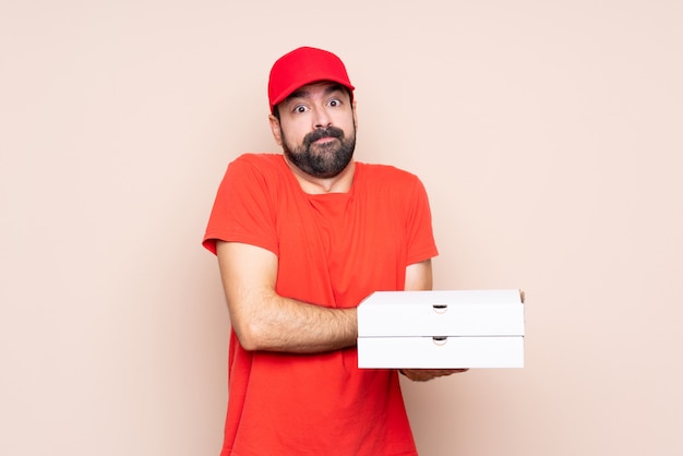 Young man holding a pizza making doubts gesture while lifting the shoulders