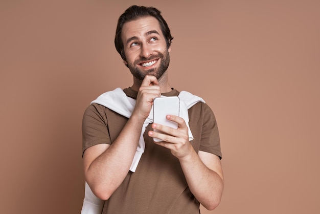 Young man holding smart phone and looking thoughtful while standing against brown background