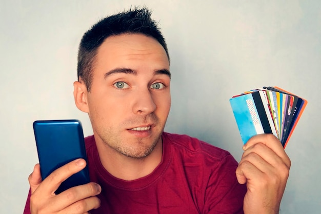 A young man holding a smartphone in one hand in the other a lot of different debit cards on a blue background choosing Bank card for payment in the online store