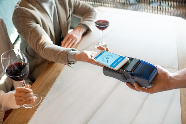 Young man holding smartphone over payment terminal while paying for dinner in restaurant while his girlfriend having glass of wine