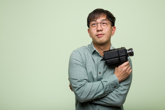 Young man holding a vintage camera smiling confident and crossing arms, looking up