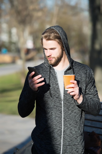 Young man holds coffee or tea paper cup and mobile or cell phone in hooded jersey sunny outdoor in park on blurred natural background morning energy blogging and internet surfing