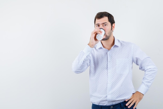 The young man is drinking coffee and putting other hand on waist on white background