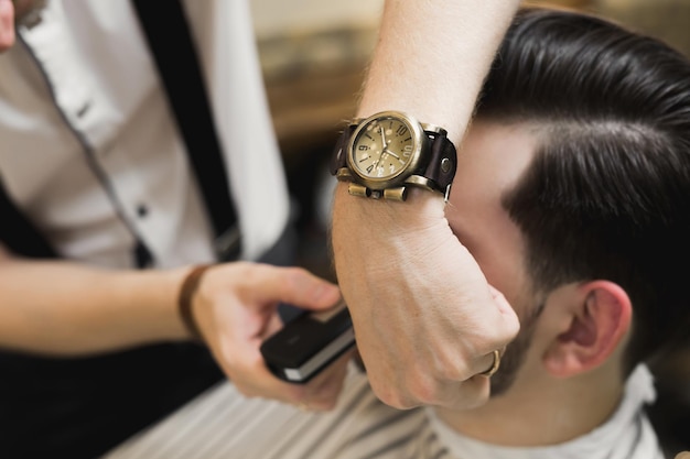 A young man is sitting in a chair in a barber shop while a barber shaves his beard