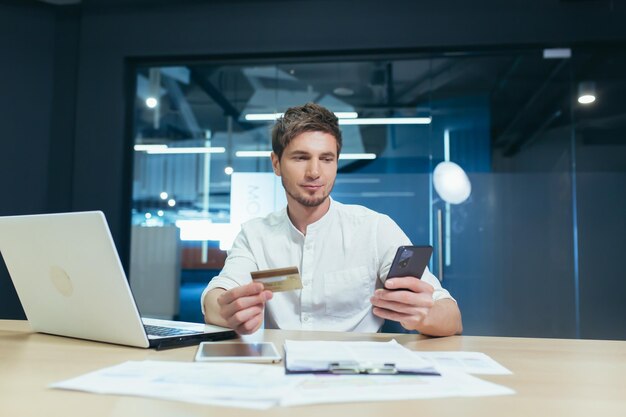 A young man is sitting in the office at his desk holding a credit card and a mobile phone working on a laptop Makes orders online pays bills