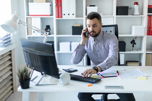 A young man is sitting in the office, talking on the phone and working on the computer.