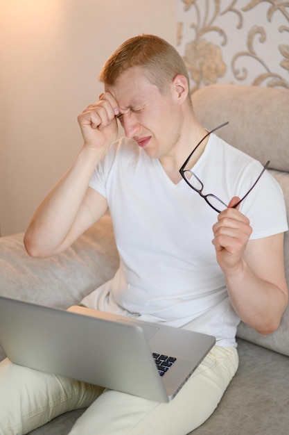 A young man is sitting on a sofa with a laptop and is experiencing a headache eye fatigue