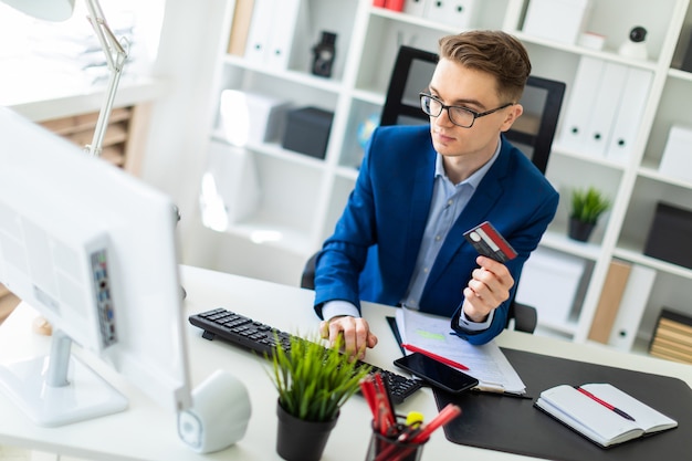 A young man is sitting at a table in the office, holding a bank card in his hand and typing  a computer.