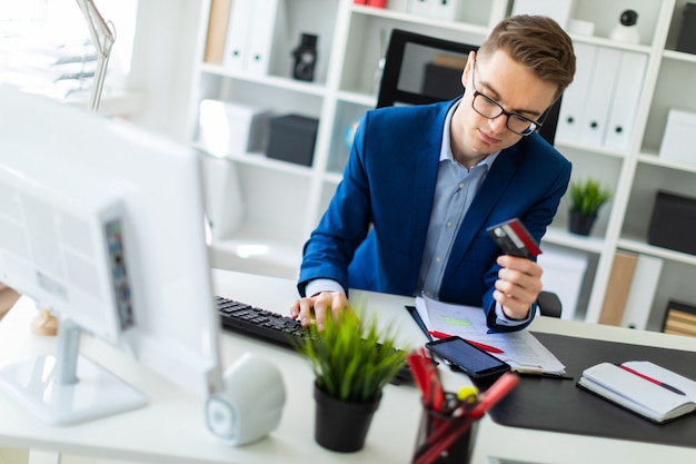 A young man is sitting at a table in the office, holding a bank card in his hand and typing  a computer.